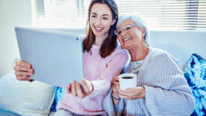 Elderly woman smiling at home while a caregiver helps her with a blanket and warm drink, illustrating winter wellness and safe homecare.
